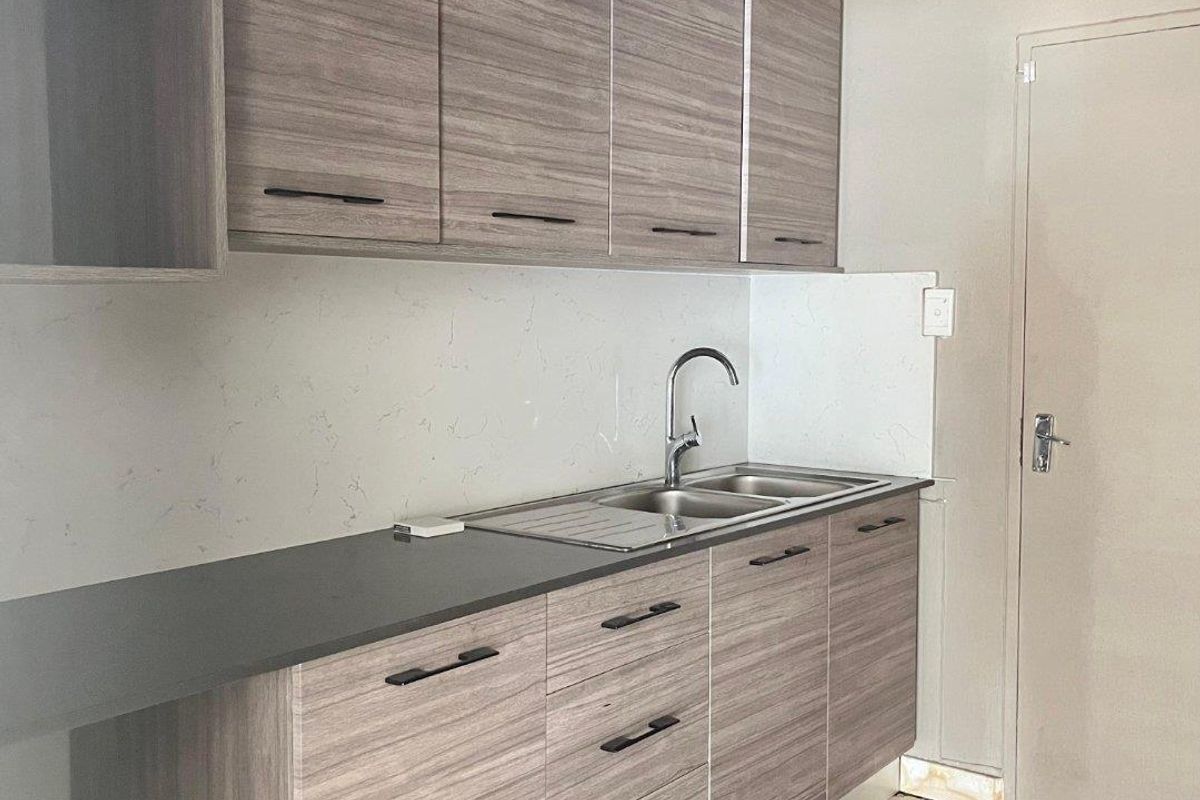 A modern, clean kitchen area featuring wood-grain laminate cabinetry, stone countertops, and a stainless steel sink with a contemporary mixer tap.