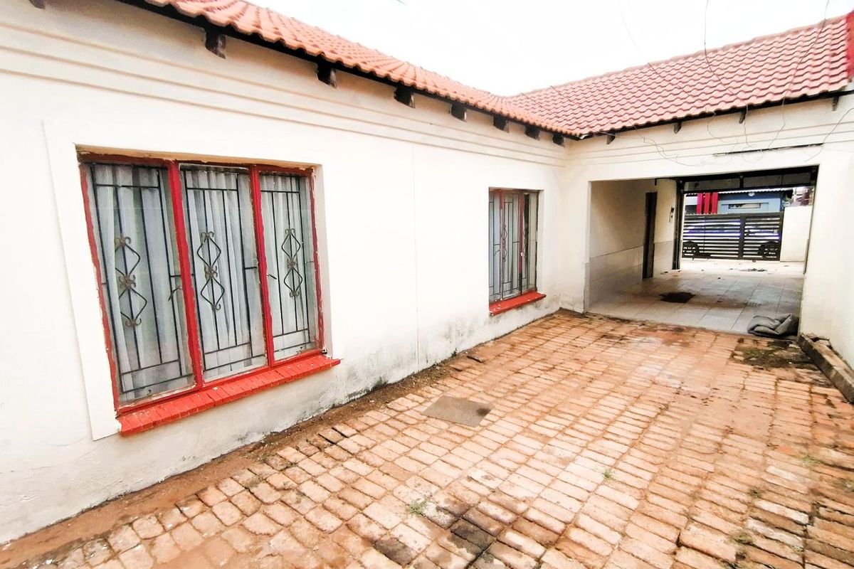 A rear exterior view of the property featuring a brick-paved courtyard, covered walkway, and tiled roof structure with security bars on windows.
