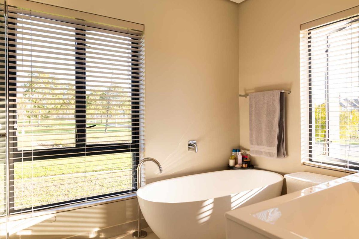 A bright, modern bathroom featuring a sleek freestanding bathtub, floor-mounted faucet, and large windows with venetian blinds allowing for ample natural light.