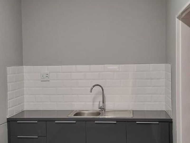 A clean, modern kitchenette featuring dark grey cabinetry, a stainless steel sink, and a white subway tile backsplash in a well-lit office space.