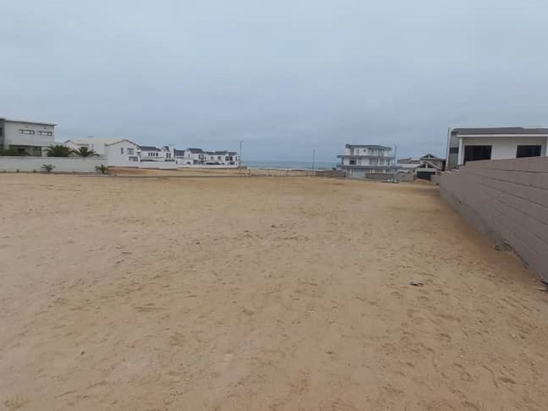 A vacant residential plot featuring sandy terrain, a boundary wall, and a paved driveway, situated in a coastal neighborhood.