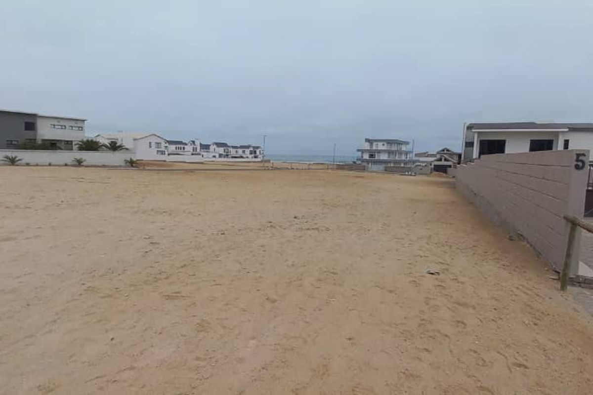 A vacant residential plot featuring sandy terrain, a boundary wall, and a paved driveway, situated in a coastal neighborhood.