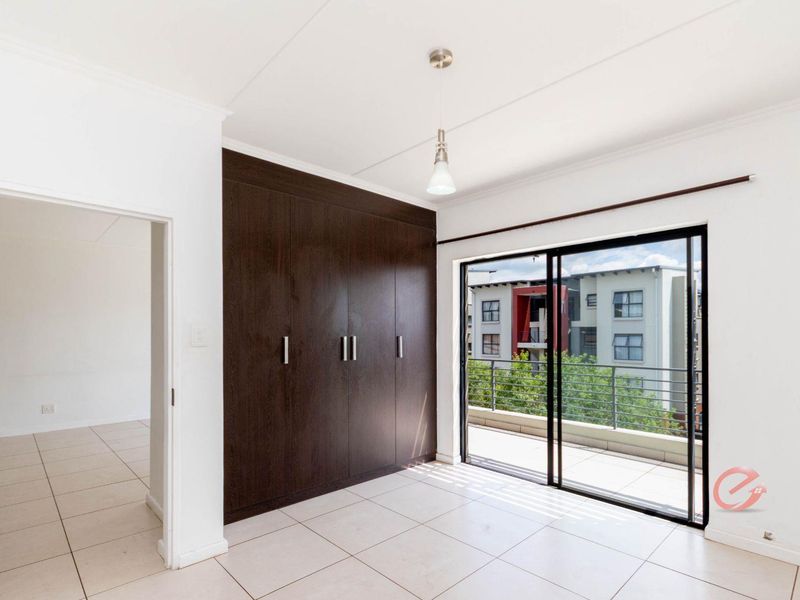 A bright, empty bedroom featuring dark wood built-in cupboards, neutral tiled flooring, and a sliding glass door leading to a private balcony.