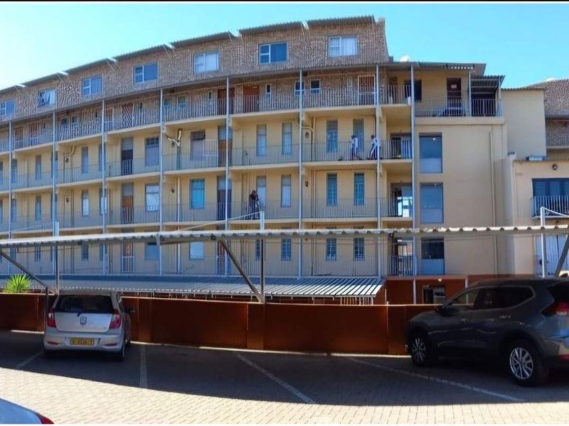 Exterior view of a multi-story apartment building featuring balconies and a covered parking area with paved driveway.