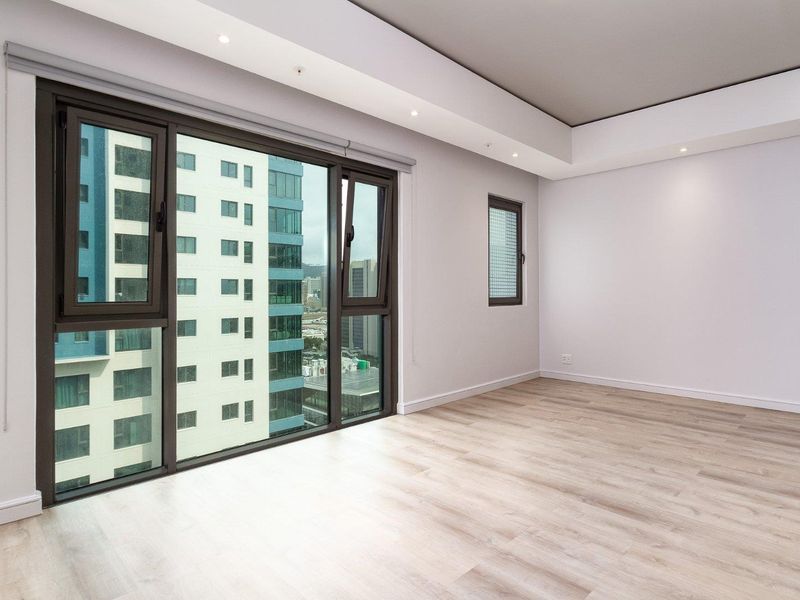 A bright, empty bedroom featuring modern laminate flooring, recessed ceiling lighting, and large aluminium-framed windows offering an urban city view.