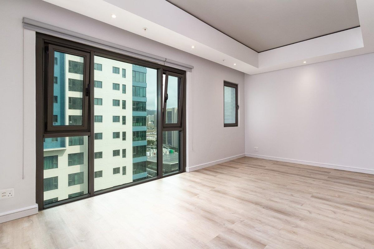A bright, empty bedroom featuring modern laminate flooring, recessed ceiling lighting, and large aluminium-framed windows offering an urban city view.
