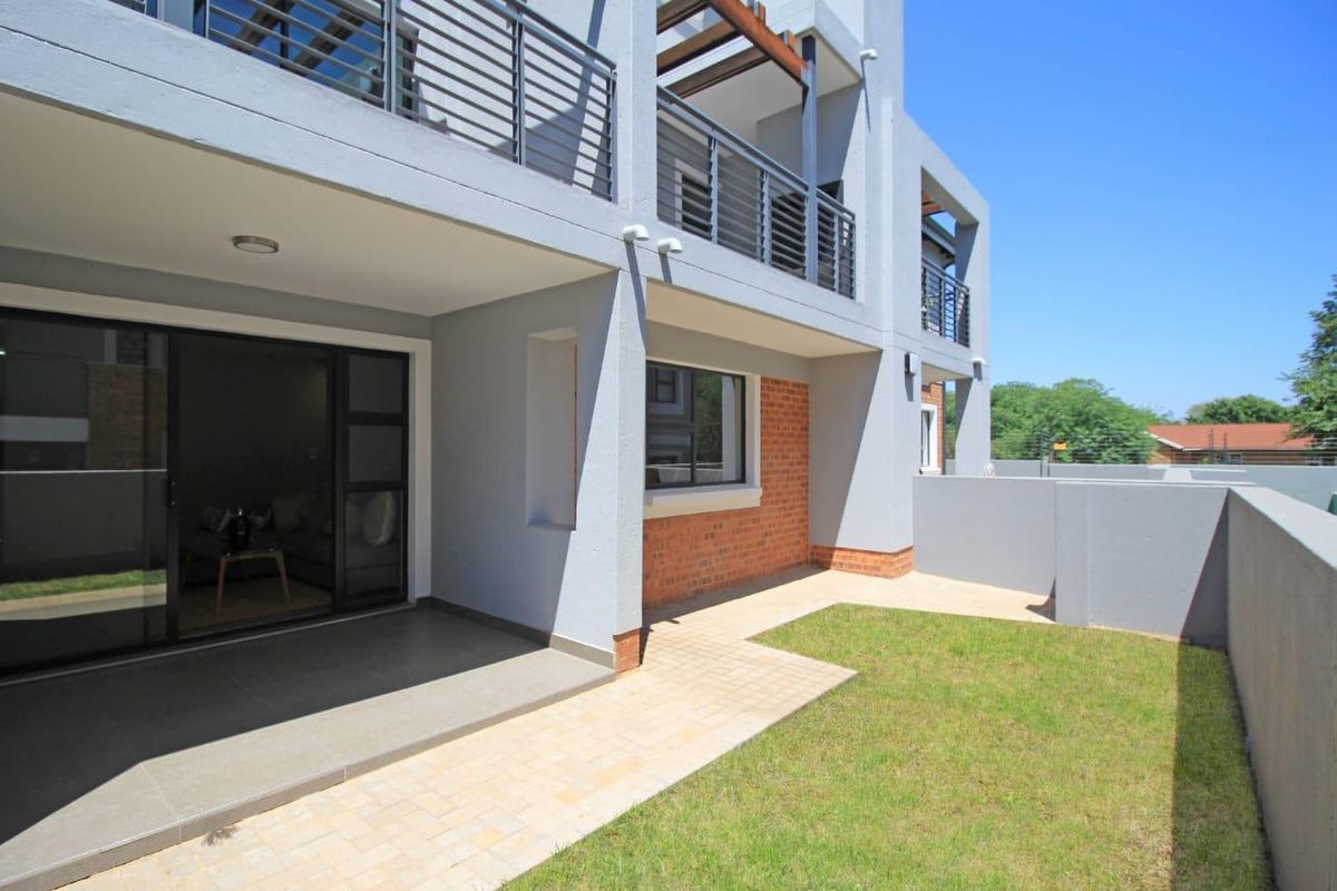 Modern apartment exterior featuring a covered patio, paved walkway, small private garden, and balcony with metal railings.