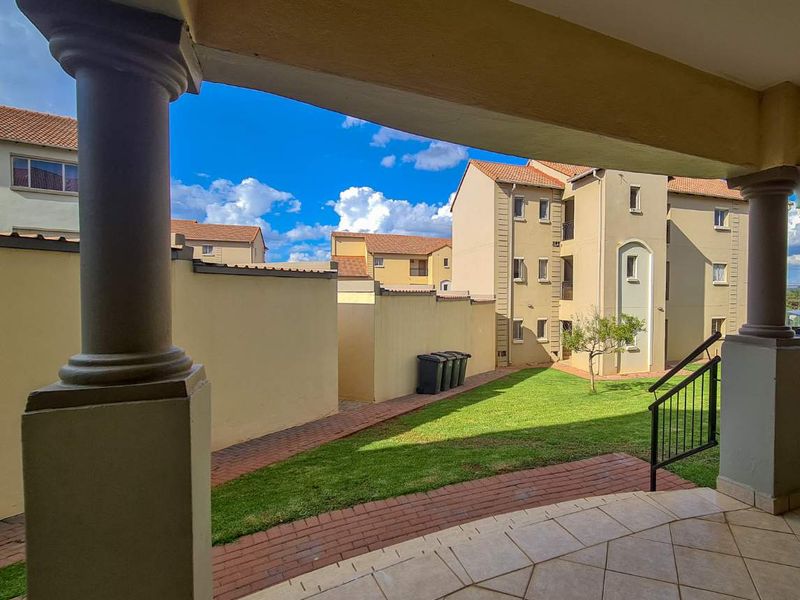 A covered patio area with tiled flooring and structural columns, overlooking a communal garden and paved walkway within the apartment complex.