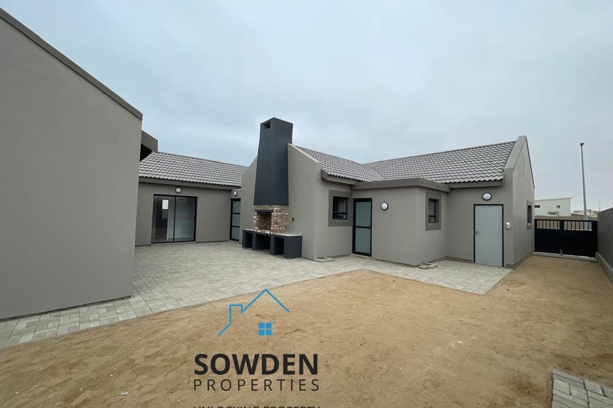 A modern rear exterior view featuring a paved patio area, a built-in outdoor fireplace, and a low-maintenance garden space.