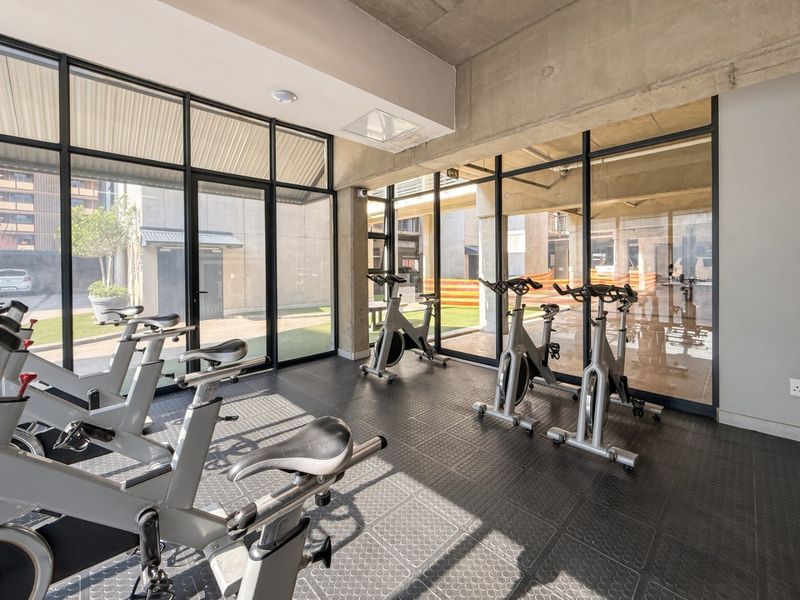 A modern, well-lit gym facility featuring durable rubber flooring, exposed concrete ceilings, and floor-to-ceiling glass walls overlooking the exterior grounds.