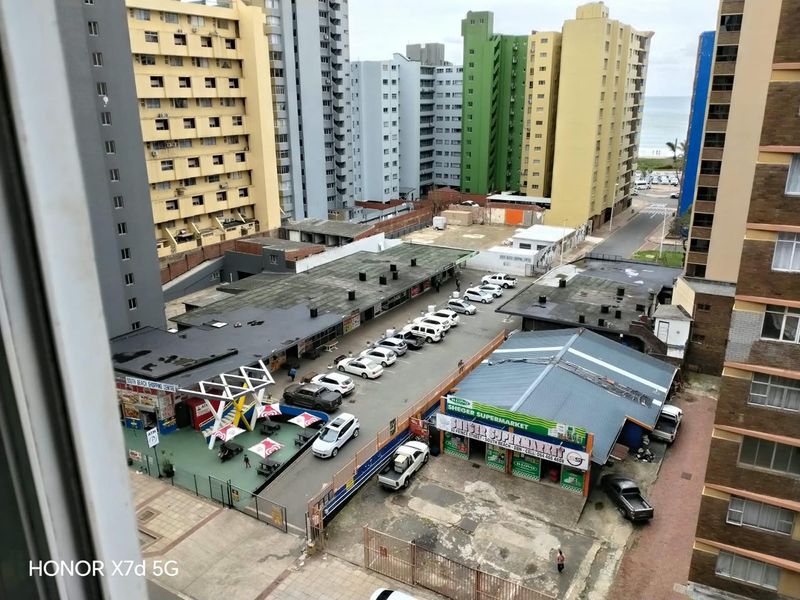 Elevated urban view overlooking a commercial parking area, surrounding apartment buildings, and a glimpse of the coastline in the distance.