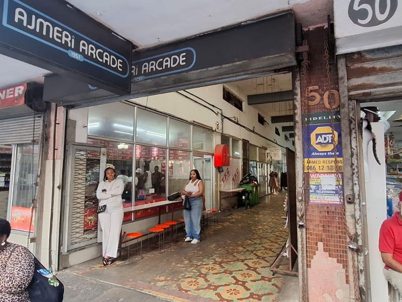 Entrance to Ajmeri Arcade featuring commercial storefronts, signage, and tiled walkway access in a busy urban setting.