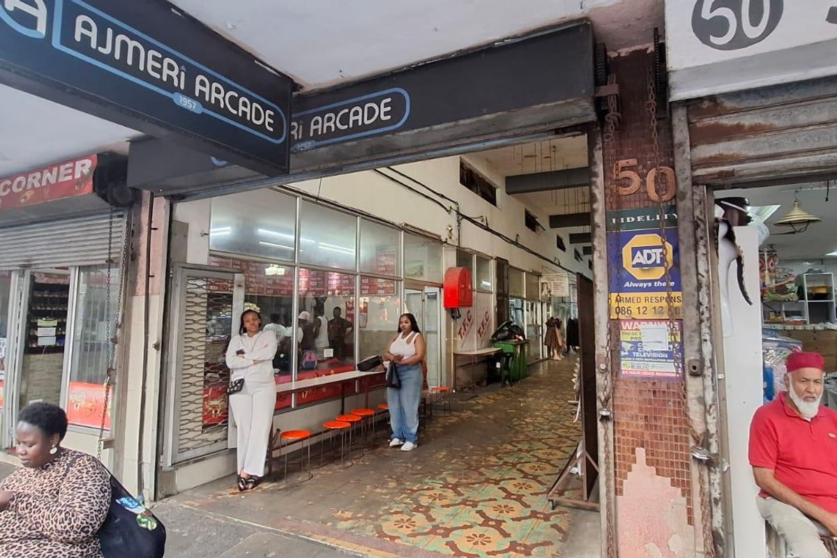 Entrance to Ajmeri Arcade featuring commercial storefronts, signage, and tiled walkway access in a busy urban setting.