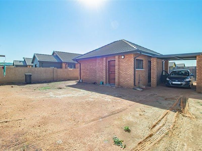 A single-story brick house featuring a tiled roof, a side carport, and a secure entrance gate, set on an unpaved plot.