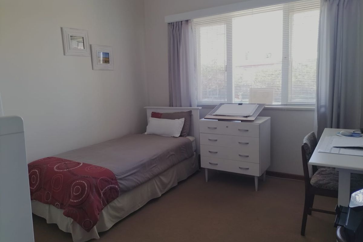 A functional bedroom featuring neutral carpeting, a large window with fitted blinds, and ample space for a single bed and study area.