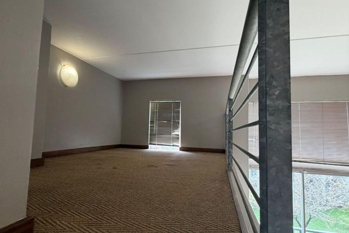 A mezzanine level bedroom area featuring carpeted flooring, a metal safety balustrade, and natural light through venetian blinds.
