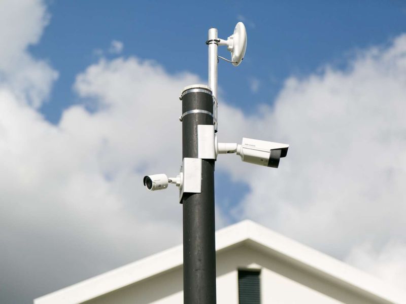A close-up view of a security pole equipped with multiple surveillance cameras and a wireless antenna, positioned against a clear sky.