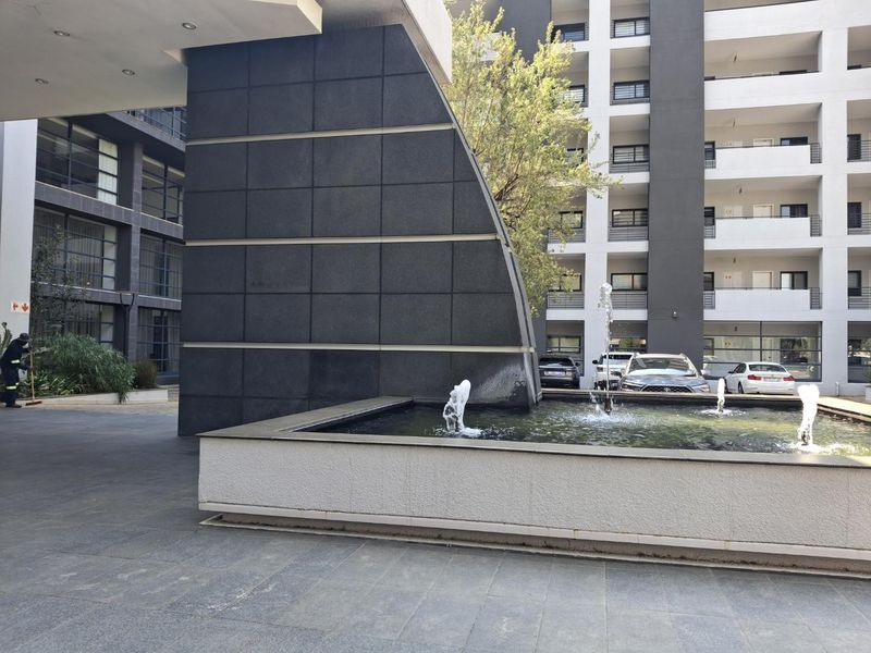 A modern apartment complex entrance featuring a decorative water fountain, paved courtyard, and contemporary architectural facade.