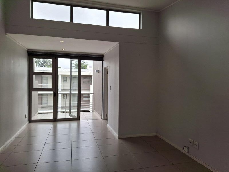 A bright, empty living area featuring large aluminium-framed glass doors leading to a balcony, tiled floors, and high-level clerestory windows for natural light.