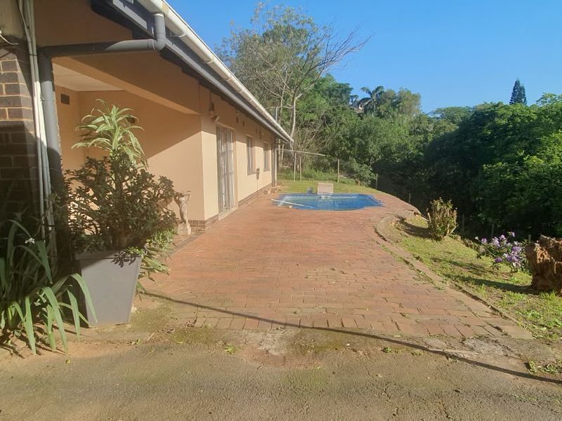 Rear exterior view featuring a brick paved patio area leading to a swimming pool, set against a backdrop of mature trees.