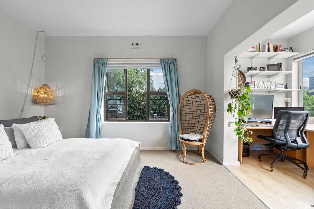 A bright bedroom featuring a carpeted sleeping area and an adjoining alcove office space with built-in shelving and large windows.