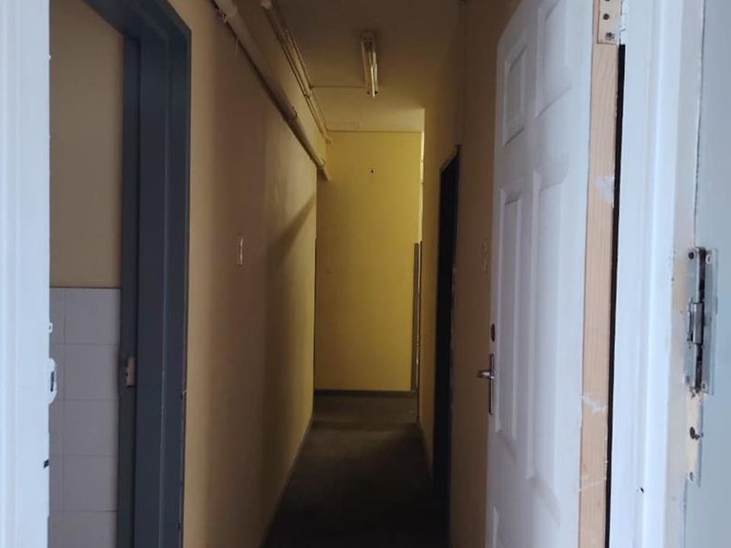 A narrow, dated interior hallway featuring exposed surface-mounted piping, fluorescent lighting, and worn wall finishes requiring maintenance and cosmetic updates.