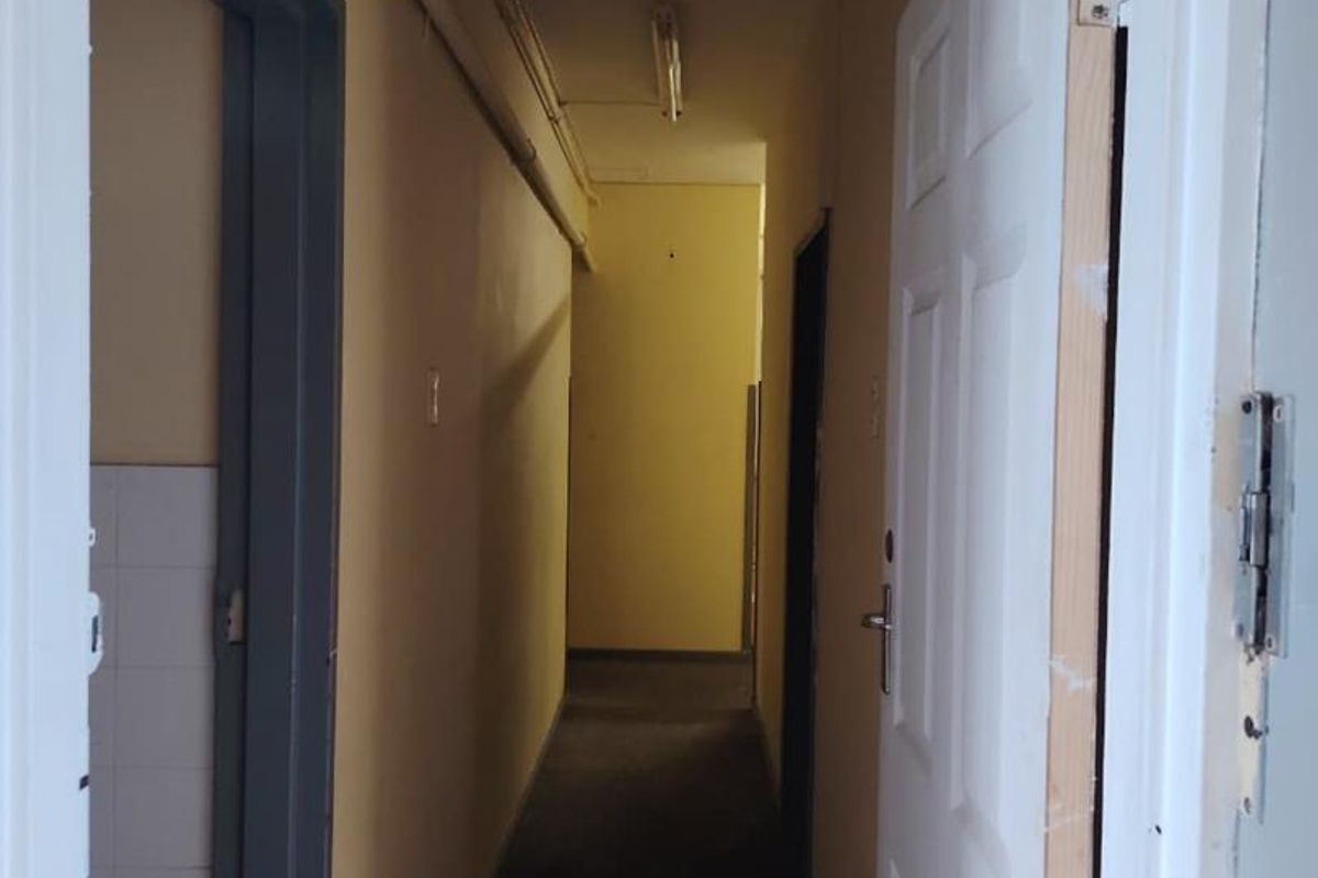 A narrow, dated interior hallway featuring exposed surface-mounted piping, fluorescent lighting, and worn wall finishes requiring maintenance and cosmetic updates.