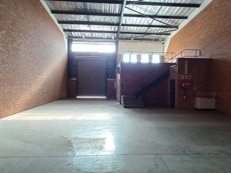 Spacious industrial warehouse unit featuring high ceilings with exposed steel trusses, concrete flooring, a roller shutter door, and a mezzanine level with staircase.