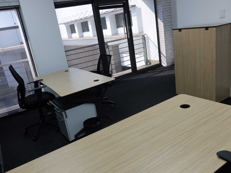 A modern office space featuring dark carpeting, exposed brick accents, and large windows providing natural light and access to a balcony.