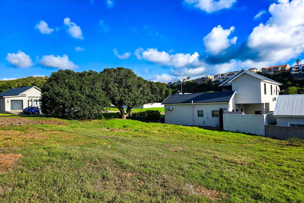 A view of a vacant residential plot featuring established trees and adjacent residential properties under a clear blue sky.