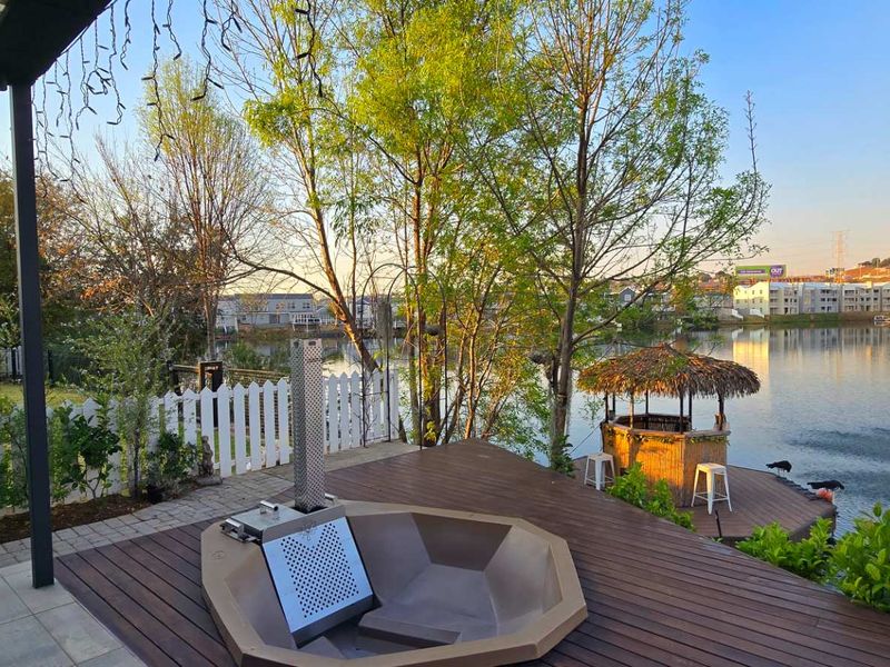 A scenic outdoor patio featuring timber decking, a built-in hot tub, and direct views over the adjacent lake and landscaped garden area.