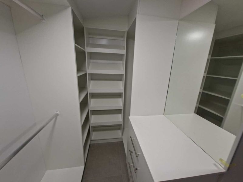 A clean, modern walk-in closet featuring white built-in cabinetry, ample shelving, hanging rails, and a built-in vanity with a large mirror.