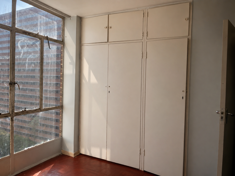 A bright, empty bedroom featuring original parquet flooring, floor-to-ceiling built-in cupboards, and a large window with an urban view.