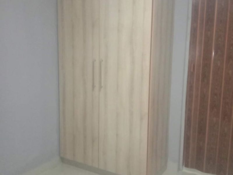 A clean, empty bedroom featuring light wood-finish built-in wardrobes, neutral grey walls, and tiled flooring.