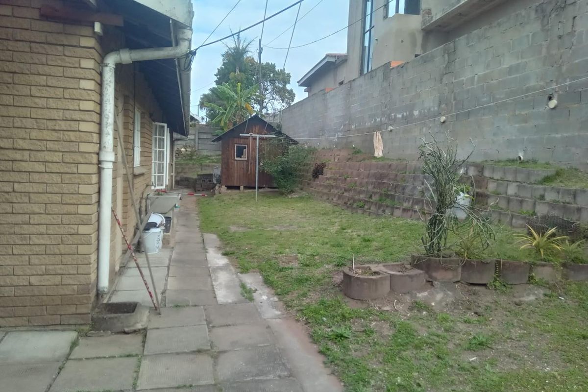 Rear exterior view showing a paved walkway, lawn area, timber shed, and tiered concrete block retaining walls along the boundary.