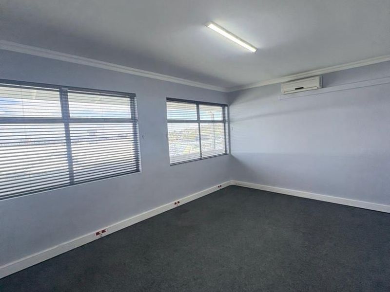 A clean, empty office space featuring grey carpeting, wall-mounted air conditioning, and large windows with fitted venetian blinds.
