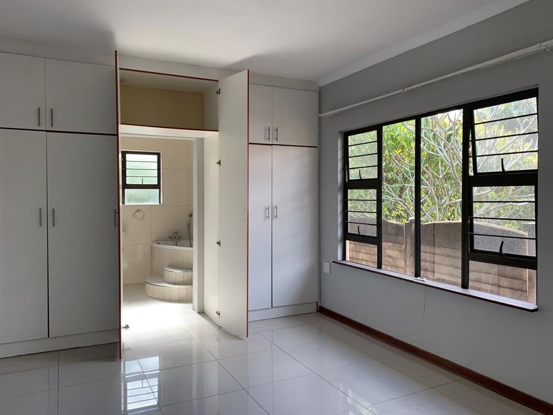 Bright bedroom featuring built-in cupboards, tiled flooring, and large windows with security bars, leading directly into an en-suite bathroom.