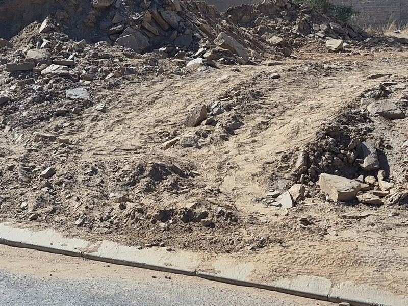 A vacant residential plot featuring uneven, undeveloped terrain with piles of rocks and soil, bordered by a concrete curb.