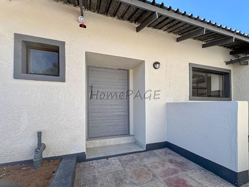 Front entrance area featuring a modern grey door, tiled threshold, and external lighting under a corrugated roof structure.