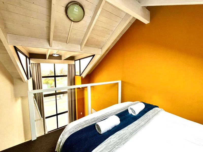 A loft-style bedroom featuring a vaulted ceiling with exposed timber rafters, a glass balustrade, and a large window overlooking the exterior.