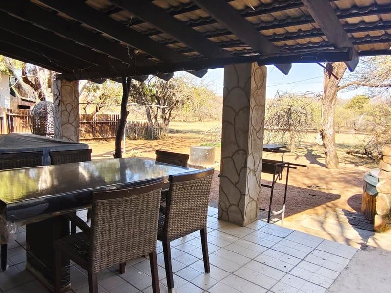 A covered outdoor patio featuring tiled flooring, stone-clad support columns, and an exposed timber beam ceiling, overlooking a natural garden area.
