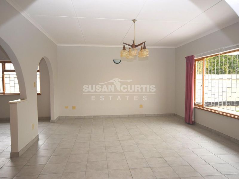 A spacious, empty living room featuring neutral tiled flooring, an arched entryway, and large windows fitted with security bars.