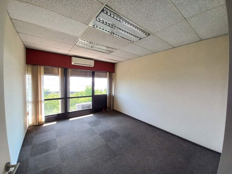 A vacant office space featuring carpeted flooring, recessed fluorescent lighting, and a wall-mounted air conditioning unit with large windows providing natural light.