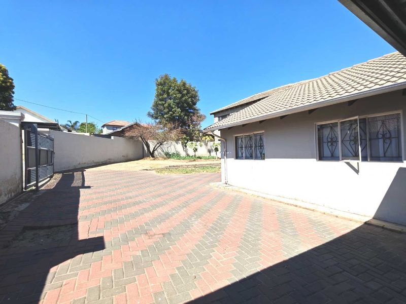 A spacious rear exterior area featuring a paved driveway, secure boundary walls, and a tiled roof dwelling with security bars on the windows.