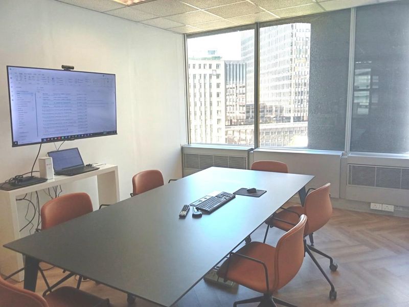 A professional office meeting room featuring parquet flooring, large windows with city views, and a wall-mounted display screen for presentations.