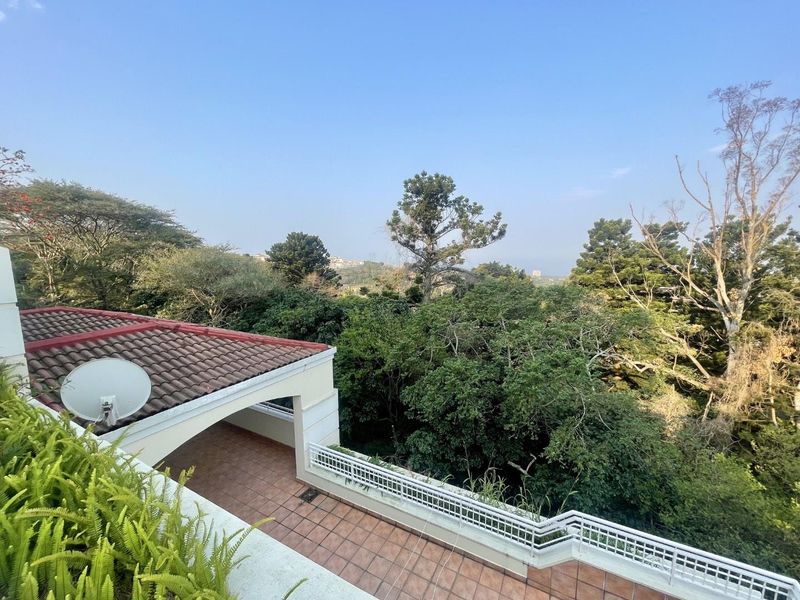A spacious covered balcony featuring tiled flooring and a metal balustrade, offering elevated views over the surrounding lush greenery.