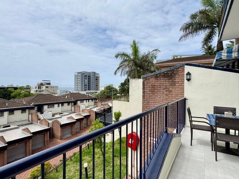 A spacious balcony featuring tiled flooring and a metal balustrade, offering elevated views of the surrounding residential complex and distant ocean.