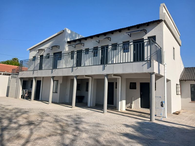 A two-story apartment building exterior featuring a paved driveway, ground-level covered parking area, and an upper-level balcony with metal railings.