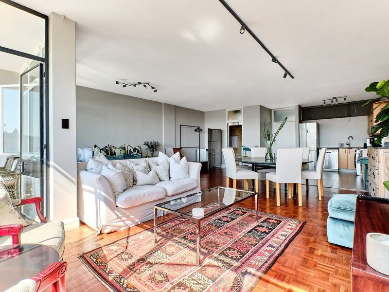 Spacious open-plan living and dining area featuring warm parquet flooring, modern track lighting, and seamless access to an adjoining balcony through large glass doors.