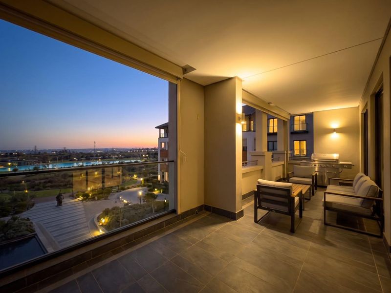 Spacious covered balcony featuring tiled flooring, a built-in gas braai, and glass balustrades, offering scenic views of the estate grounds at dusk.